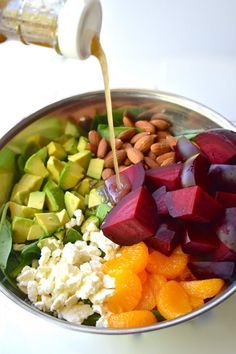 someone pouring dressing into a bowl filled with vegetables and fruits, including beets, avocado, carrots, almonds, cucumber, and other ingredients