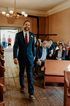 a man in a suit and tie walking down the aisle with people sitting on chairs behind him