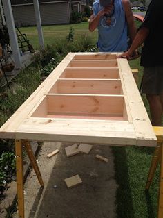 two men are working on an unfinished table in the yard, with one man standing next to it