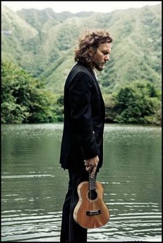 a man holding a guitar standing in front of a body of water