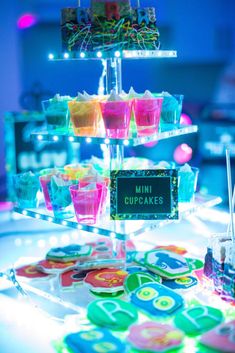 colorful cupcakes are displayed on a clear cake stand with lights in the background