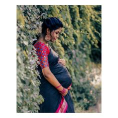 a pregnant woman in a black and red sari standing next to some bushes with her belly exposed