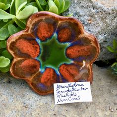 there is a small flower shaped bowl on the ground next to some plants and a note