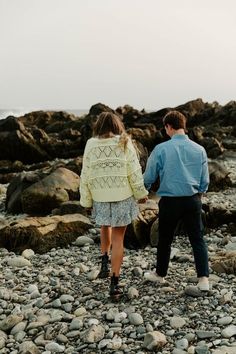 a man and woman holding hands walking on rocks near the ocean with water in the background