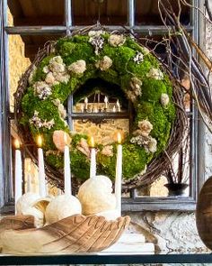 a wreath with white candles and flowers on top of a mantle in front of a window