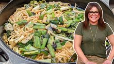 a woman standing in front of a skillet filled with pasta and asparagus