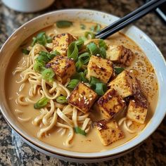 a bowl of noodles and tofu with chopsticks in it on a table