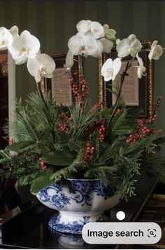 white flowers in a blue and white vase on a table with red berries, pine cones and greenery