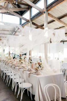 a long table with white chairs and tables covered in sheer fabric, is set up for an event