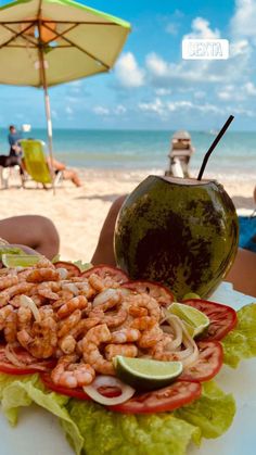 a plate with shrimp, lettuce and tomatoes on it next to an umbrella at the beach