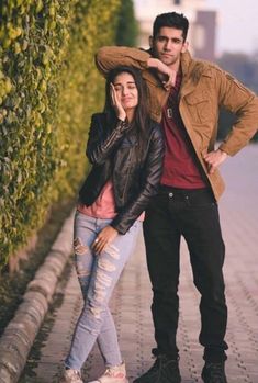 a man and woman standing next to each other on a sidewalk with hedges behind them