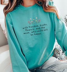 a woman sitting on a couch wearing a blue sweatshirt with flowers and the words,'with freedom, books, flowers, and not moon, who could not be happy