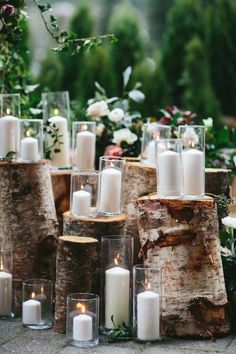 white candles are placed in glass vases next to wooden logs with greenery and flowers