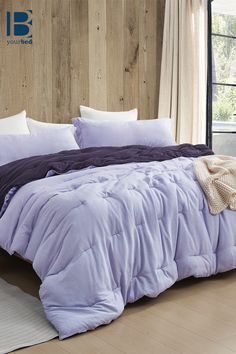 a bed with purple comforter and pillows in a room next to a wooden wall