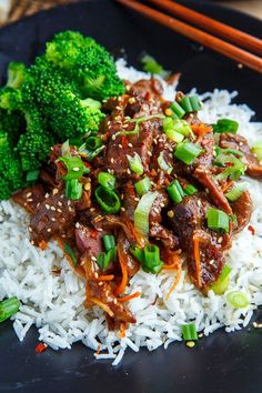 a black plate topped with white rice and meat covered in sauce next to broccoli