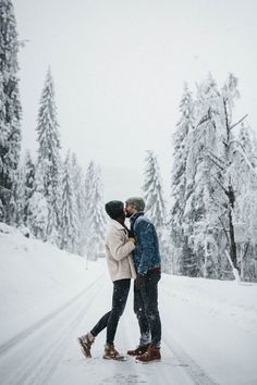 a man and woman standing in the middle of a snow covered road with trees behind them