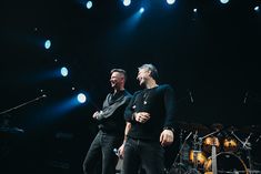 two men standing on stage with microphones in front of them and lights behind them