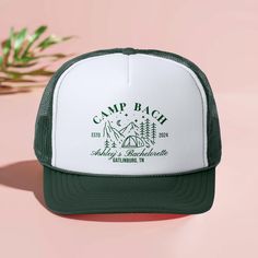 the camp beach trucker hat is shown on a pink surface with a plant in the background