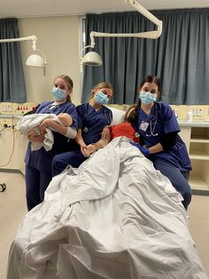 three women in scrubs and masks stand around a hospital bed