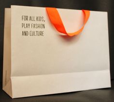 a white paper bag with an orange ribbon on the handle that says for all kids, play fashion and culture