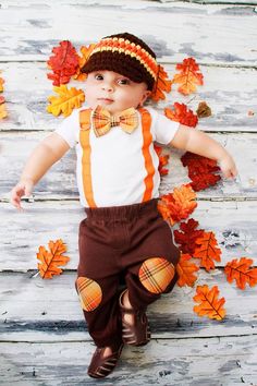 a baby wearing a hat and bow tie laying on the ground with autumn leaves around him