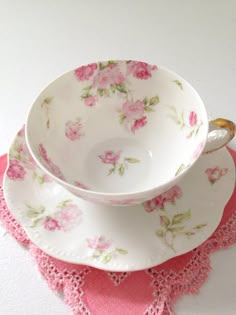 two tea cups and saucers with pink flowers on them sitting on a doily