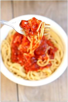 a fork full of spaghetti with sauce on it in a white bowl over a wooden table