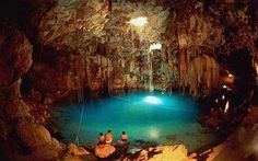 two people sitting on rocks in front of a blue pool surrounded by cave like formations