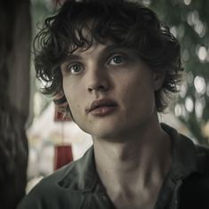 a young man with curly hair stares into the distance while standing in front of a tree