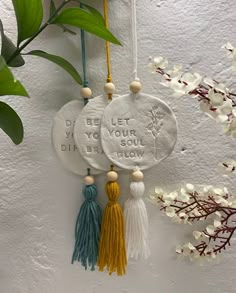 three tasseled ornaments hanging from a wall with flowers in the background and words written on them