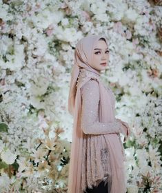 a woman wearing a hijab standing in front of a flowery wall with white and pink flowers