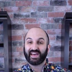 a man with a beard and blue shirt in front of a brick wall, looking at the camera