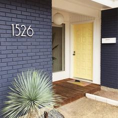 a yellow door is on the side of a blue brick house with a plant in front