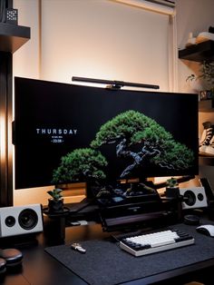 a computer desk with a monitor, keyboard and speakers in front of the monitor is a bonsai tree