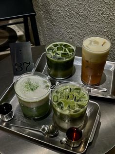 three different types of drinks on a metal tray with spoons and cups next to each other