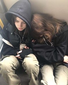 two people sitting on a bus looking at their cell phones and one person is wearing a hoodie