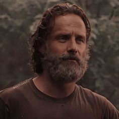 a man with long hair and beard wearing a brown t - shirt in front of trees