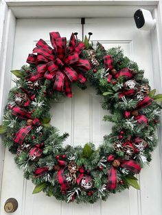 a christmas wreath hanging on the front door with red and black plaid bow, pine cones, evergreen leaves and bells