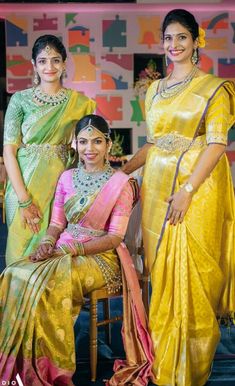 three women in yellow and pink saris posing for the camera with one woman sitting on a chair