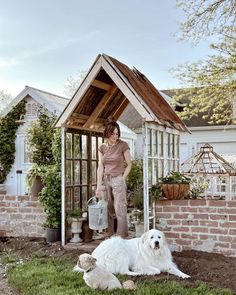 a woman standing next to a white dog in front of a brick wall and shed