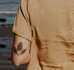 a man with a pine cone tattoo on his left arm next to the ocean and sand