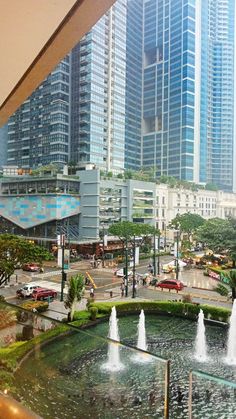 a view of a city from inside a building with fountains in the foreground and cars on the street