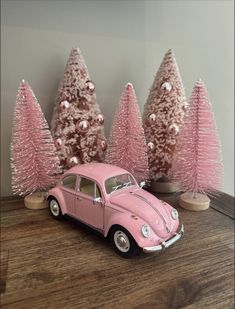 a pink vw bug parked in front of christmas trees on a wooden table next to a wall