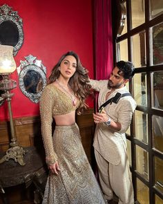 a man standing next to a woman in a white and gold dress on a red wall