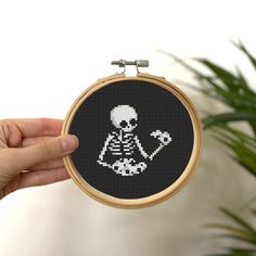 a person holding up a cross stitch skeleton in front of a potted green plant