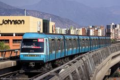 a blue train traveling over a bridge in front of tall buildings and mountains behind it