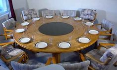 a round wooden table surrounded by chairs and place settings with plates on the center, in front of a window