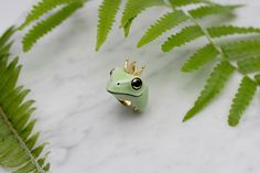 a frog with a crown on it's head sitting next to a fern leaf