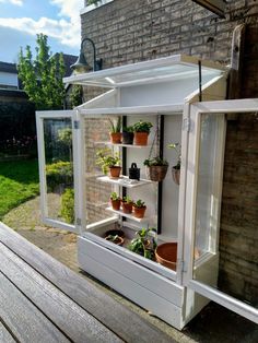 a white greenhouse with potted plants in it on a wooden deck next to a brick building