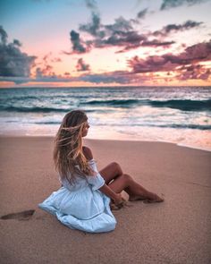 a woman sitting on top of a sandy beach next to the ocean at sunset with her hair blowing in the wind